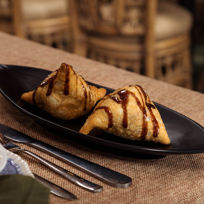 Two golden-brown fried pastries topped with tamarind chutney drizzle, served on a black plate on a woven beige tablecloth with cutlery and a softly blurred restaurant background.